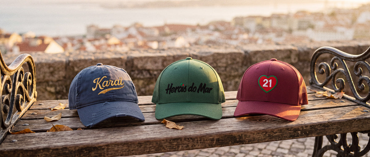 Trois casquettes brodées TifoArt sur un banc d'un miradouro de Lisbonne au coucher du soleil : Karaï délavée marine, Héróis do Mar verte et 21 cranberry