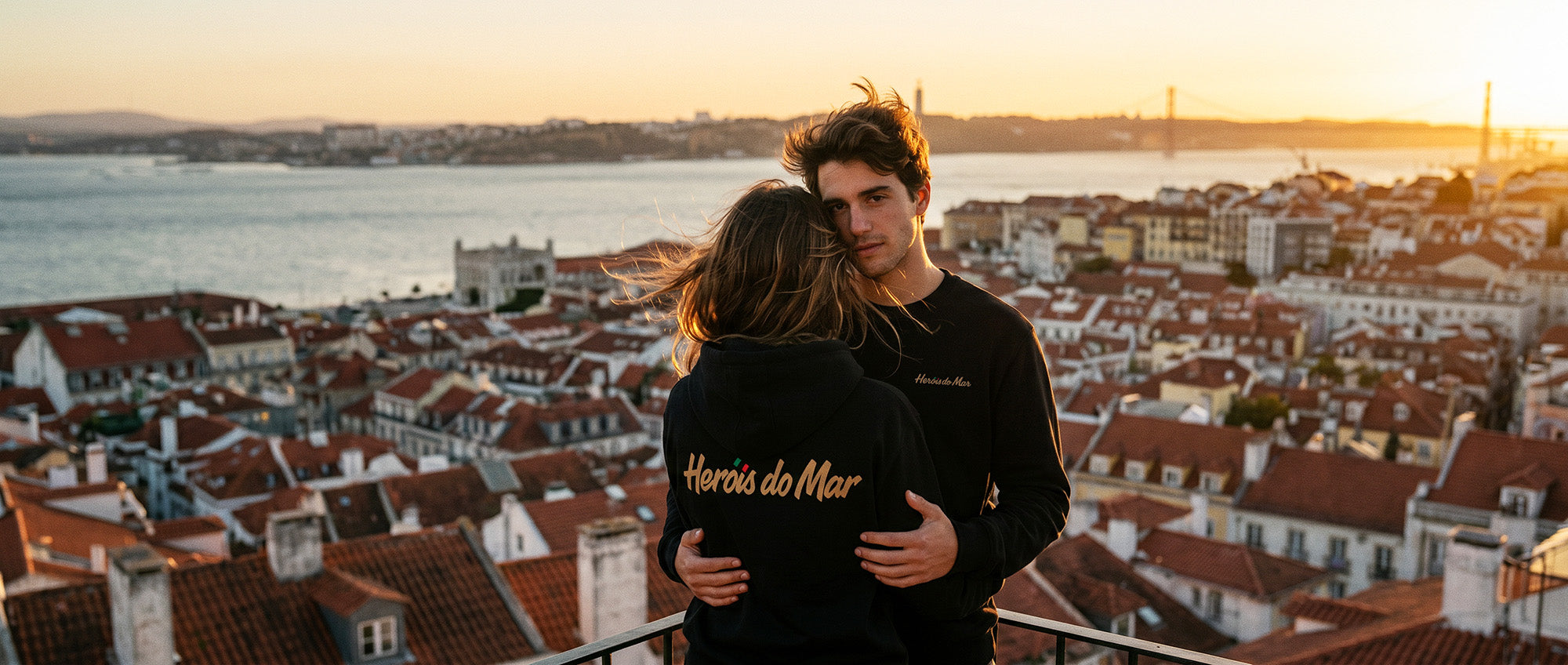 Couple de la diaspora portugaise enlacé à un belvédère de Lisbonne au coucher du soleil, sweat Heróis do Mar TifoArt et pont du 25 Avril en fond