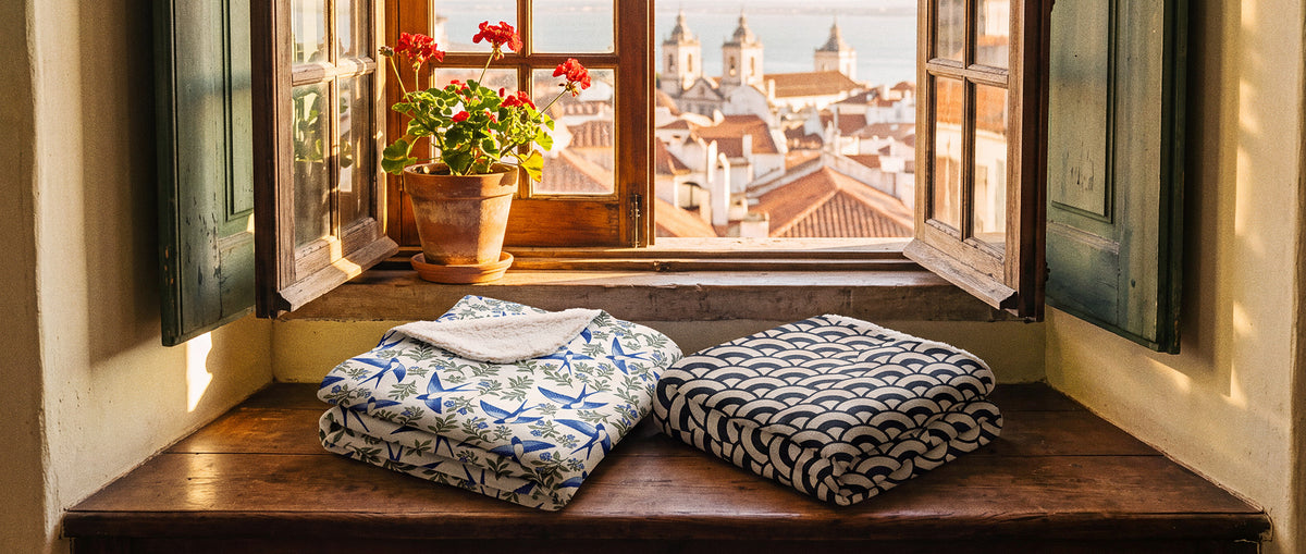 Deux plaids sherpa TifoArt pliés sur un banc de fenêtre à Lisbonne, motifs hirondelles et calçada, vue sur Alfama